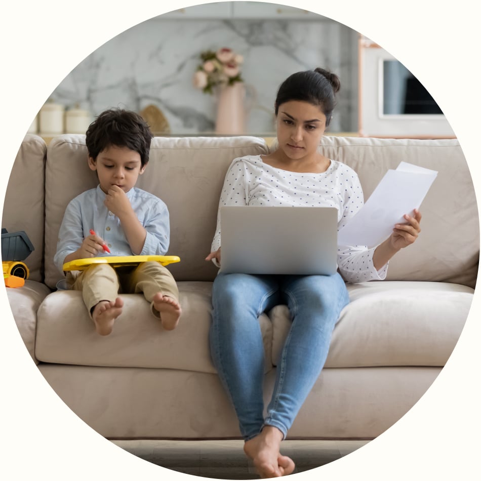 young mother sitting on sofa at home paying bills on a laptop computer while her child sits next to her