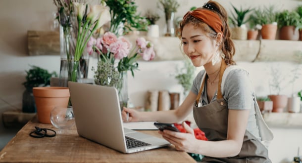 Asian female florist, owner of small business flower shop, using smartphone while working on laptop