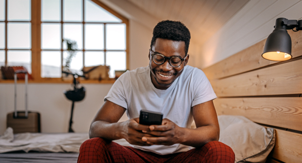smiling young man with eyewear in casual clothing checking his smart phone sitting on bed