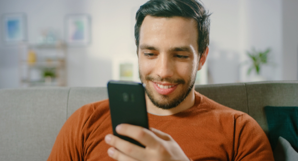 Happy Young Man Uses Smartphone while Sitting on a Sofa at Home