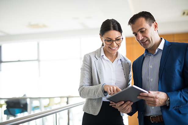 Two mixed-age business people reviewing data on digital tablet in office hallway.