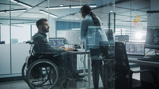 Data security specialist in a wheelchair talks with colleague in office while working on computer.