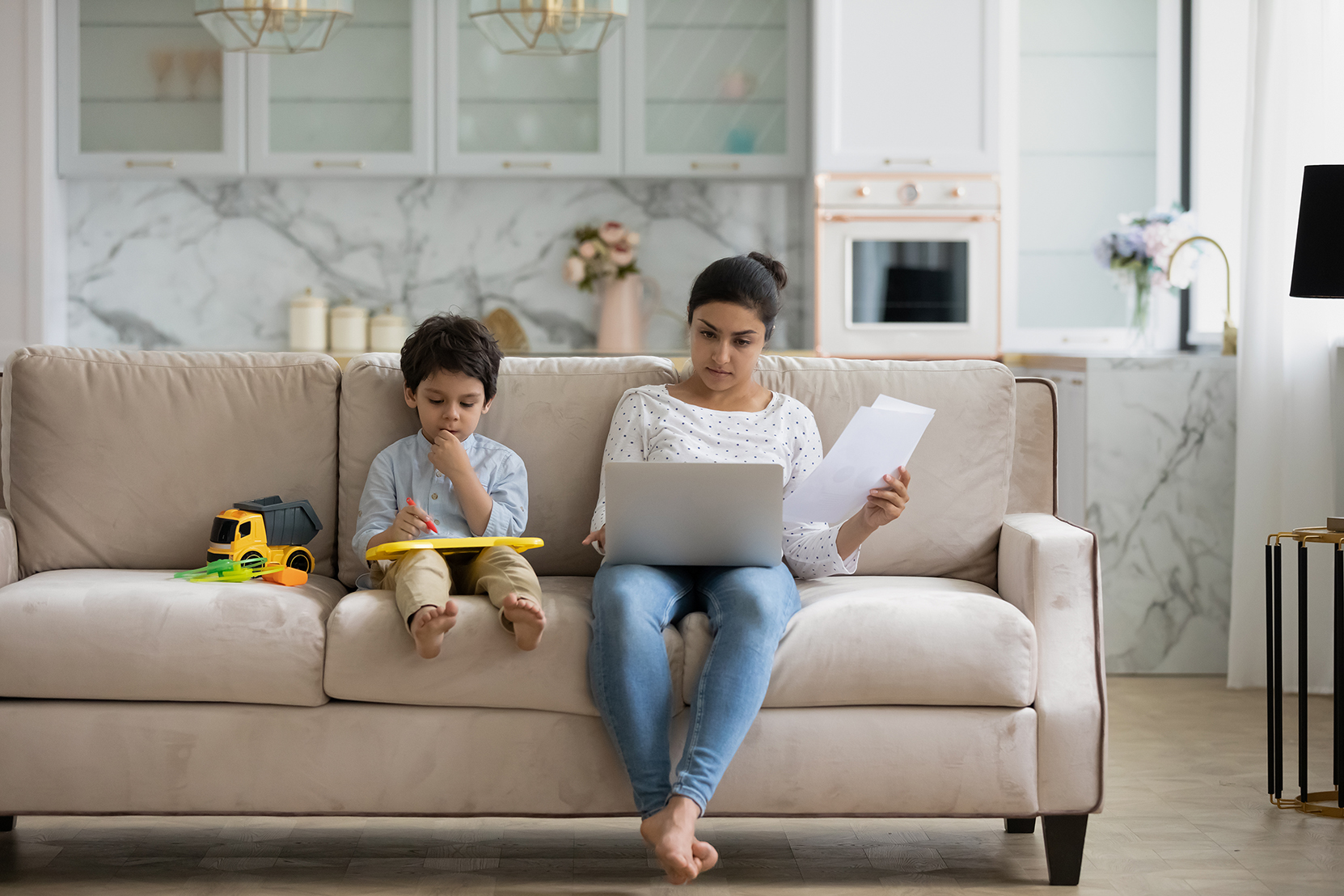 young mother sitting on sofa at home paying bills on a laptop computer while her child sits next to her